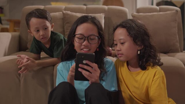 Three young siblings enjoy watching a smartphone together on a sofa in the living room, capturing children's screen time, family togetherness, home entertainment, and modern family lifestyle.