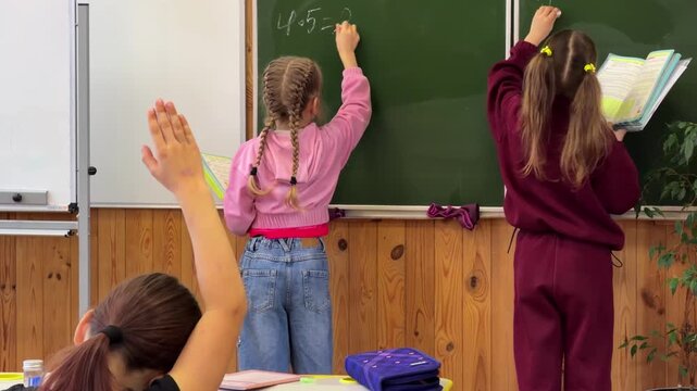 Students writing math equations on a green classroom blackboard while peers raise hands in a primary school setting to participate in an interactive lesson about academic learning