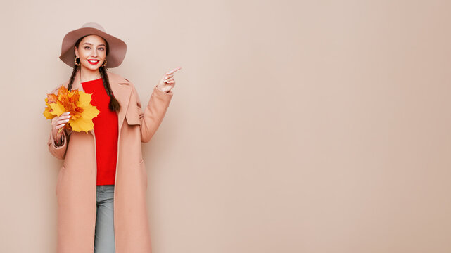 A beautiful young woman in an autumn fashionable outfit with shopping bags and round hat pointing aside at free space over beige background