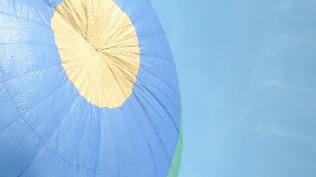 The curved top of a blue and yellow hot air balloon begin to inflate smooth waving in the wind against a bright blue sky, hot air balloon