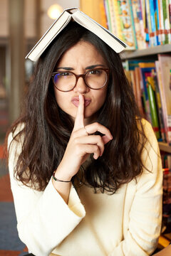 Portrait of young woman with book on top of head asking for silence at the library