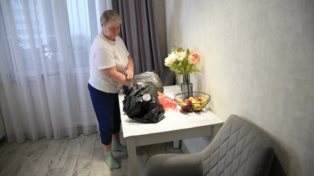 Woman folds plastic bags in an indoor setting, organizing household items on a white table next to a window with curtains and a decorative flower arrangement.