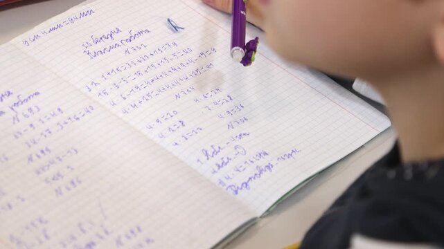 A student writing math problems in a notebook at a classroom desk, depicting focus and active learning during a primary school lesson to illustrate academic growth and development