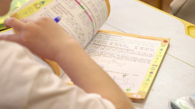 A primary student works on math problems inside a workbook at a desk, representing academic progress and focused learning in a school classroom setting during a lesson on arithmetic