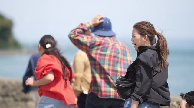 Group strolls seaside with sea breeze in background. Man adjusts hat while others walk beside him. Woman in black coat walks with ponytail down. Scene feels relaxed and casual near ocean