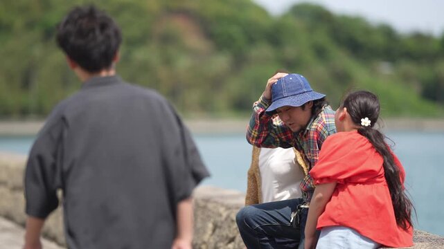 Sunlit seaside, a woman and child share quiet moments. Woman in gold drape gazes toward horizon, calm and thoughtful. Child in red shirt adjusts cap, eyes lowered, shy and curious