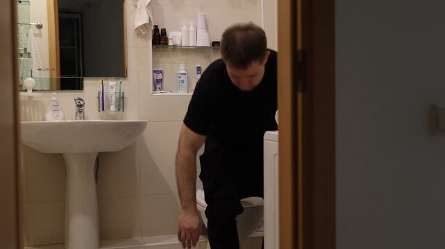 Young man leaning over and scrubbing a white toilet bowl with a yellow sponge in tiled home bathroom