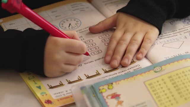 A student writing in a workbook with a red pen to solve math exercises while seated at a desk, conveying academic learning, diligence, and elementary education in a classroom setting