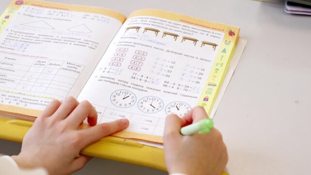 A young student completes math exercises in an open workbook while holding a highlighter at a bright desk, illustrating academic focus and primary school learning in the classroom