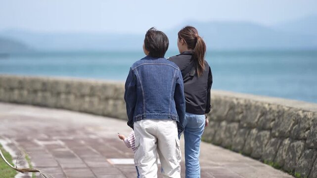 Family strolls by the sea's edge. They walk side by side, hands linked. The ocean whispers, distant and wide. Mountains fade softly in the haze. This moment feels warm and carefree