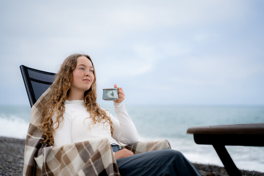 Woman enjoying a hot drink by the ocean from a lounge chair. Peaceful seaside relaxation, slow living and outdoor wellbeing concept.