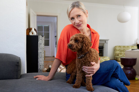 A woman sits on a sofa holding her sweet toy poodle at home
