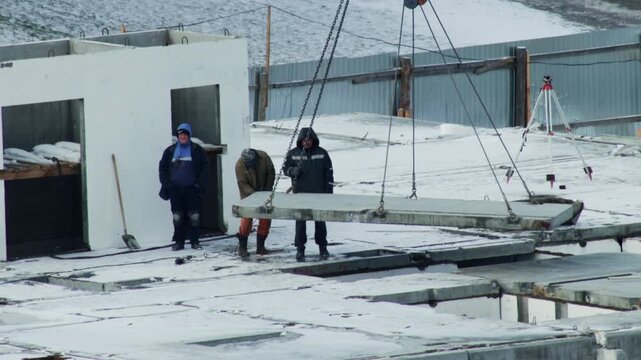 Construction workers on building site carefully lower large precast concrete slab into position using industrial crane, covered in snow on cold winter day, construction of new structure