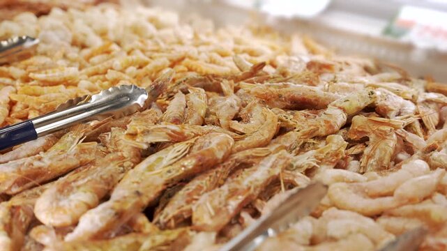 Rows of frozen prawns and peeled shrimp lie on an icy seafood display with metal tongs ready for customers to serve themselves.