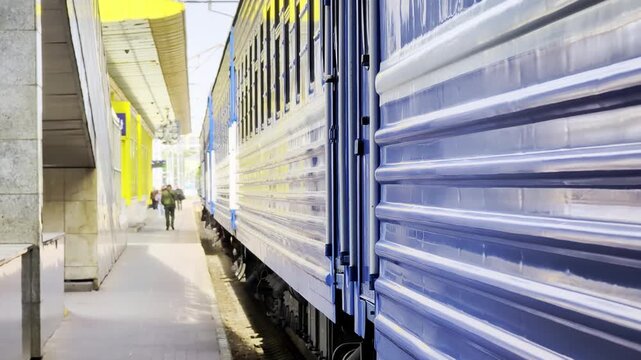 Side view of blue train car with yellow stripe stopped at station platform, showing textured exterior, multiple windows, metal components, railway track
