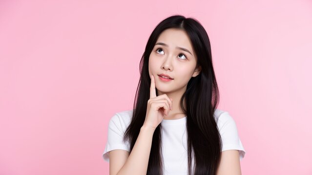 Pensive Young Woman: A young woman with long dark hair, wearing a white t-shirt, rests her chin on her hand, gazing upwards with a thoughtful expression against a soft pink background.