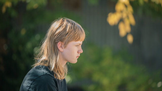 Young person with blond mullet hairstyle looking thoughtfully into the distance outdoors