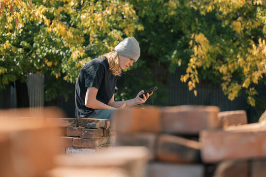 Young person in beanie looking at phone outdoors sitting among piles of bricks