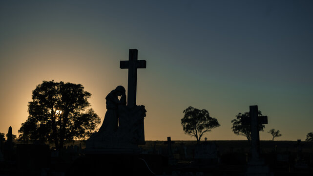 Silhouette of a weeping angel statue and cross at sunrise in a cemetery