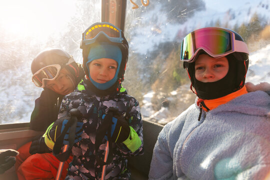 Children of different ages ride ski gondola in winter gear, with bored teenager leaning by window while younger skiers look cheerful. Warm sunlight, helmets, goggles and poles frame family ski trip