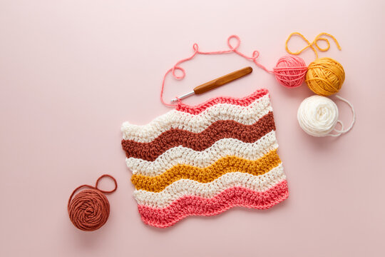Colorful crochet project with yarn balls and crochet hook on pastel pink background. DIY crochet project with colorful wool and hook, flat lay.