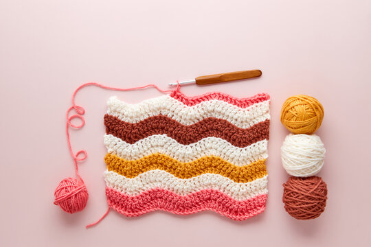 Colorful crochet project with yarn balls and crochet hook on pastel pink background. DIY crochet project with colorful wool and hook, flat lay.