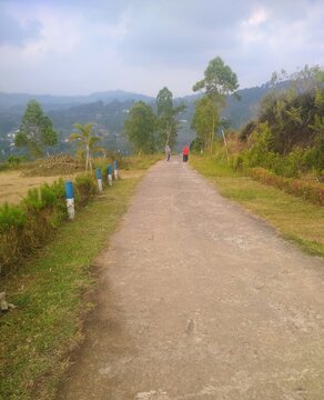 Malang, Jawa Timur, Indonesia - 15 July 2023: A peaceful rural concrete road leading towards green hills and Hill Nirwana. Two people walking in the distance with trees, grass, and village scenery.