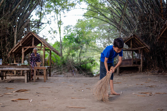 Young boy sweeps dry ground with straw broom beside rustic bamboo huts while elderly woman stands nearby in shaded rural village yard, surrounded by tall arching trees and soft natural light