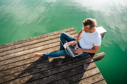 Freelancer working on laptop sitting at pier over lake