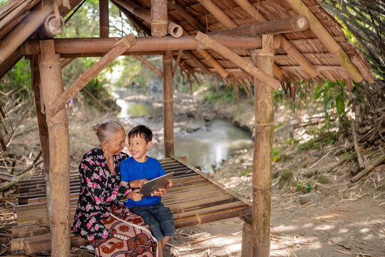 Elderly woman and young boy sit together on bamboo platform under rustic shelter beside quiet stream, smiling while sharing tablet in sunlit rural setting with natural textures, gentle greenery, and