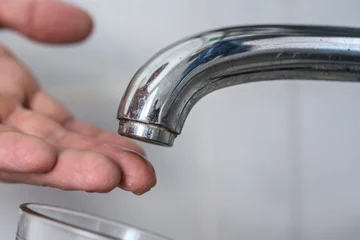 Adult hand held under chrome faucet spout with corroded aerator and mineral deposits, dry plumbing fixture showing no water flow, soft neutral background, natural skin tones and detailed metal surface © Denys Kurbatov