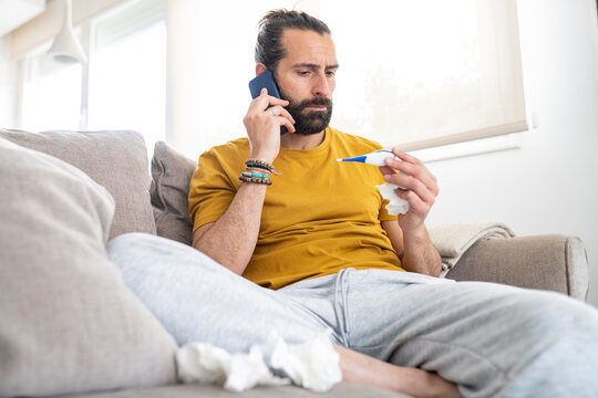 Worried man talking on smart phone while looking at thermometer at home