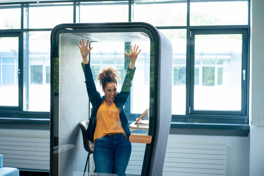 Cheerful businesswoman with hands raised inside soundproof cabin at office