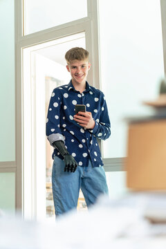 Smiling teenage boy with amputated arm holding smart phone by door