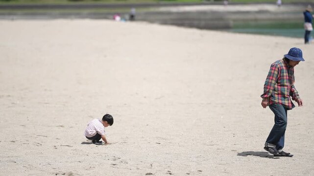 Sunlight glints off the wide, empty beach. Child kneels, digging sand with curious delight. An older man walks by, relaxed, unbothered. Soft shadows stretch across sand and water