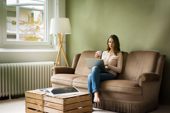 Young woman sitting on couch with cup of coffee using laptop