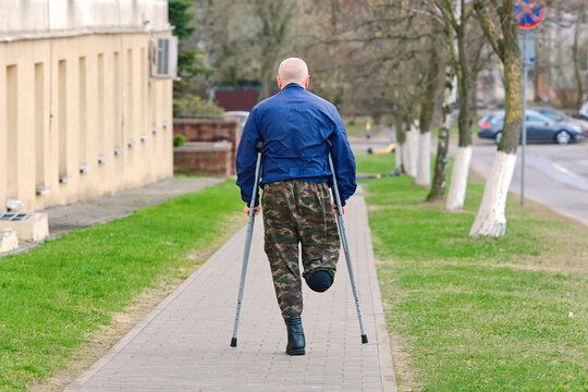 Elderly unrecognizable person with an amputated leg walking with crutches along paved city sidewalk wearing camouflage trousers, blue jacket on clear day, Physical challenges and mobility limitations