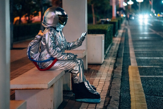Spaceman sitting on bench at a bus stop at night holding cell phone