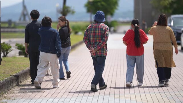 Group strolls along seaside path. People walk casually, enjoying sunny day. Background shows distant tower and calm waters. Scene captures relaxed, everyday coastal moment. Ideal for travel, lifestyle
