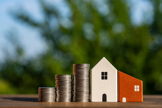 Three increasing stacks of silver coins stand next small wooden toy houses on wooden surface against blurry green nature background, symbolizing property savings, closeup