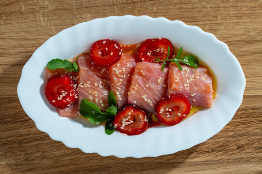 Salmon carpaccio with strawberries and sesame seeds, closeup. Gourmet dish of sliced raw salmon sashimi or carpaccio paired with fresh strawberries, sesame seeds and green arugula on white oval plate