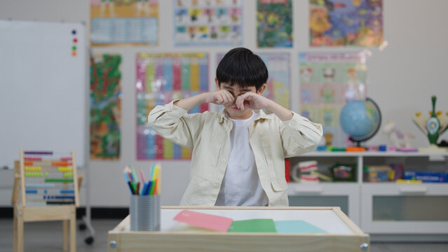 Young Asian Schoolboy Rubbing His Eyes While Sitting at Classroom Desk