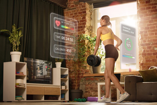 Woman performing dumbbell row with virtual workout dashboard at home. Concept of digital fitness coaching, exercise tracking, smart home gym and performance monitoring.