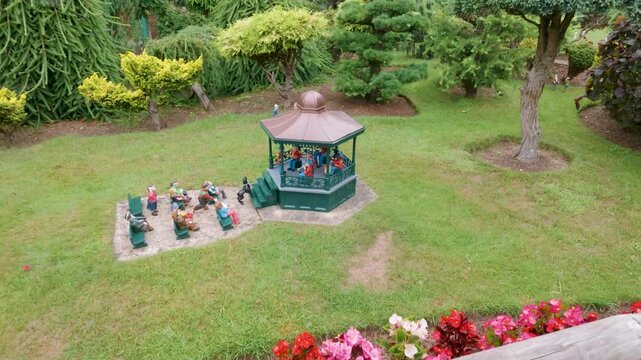 Model village bandstand in Godshill, Isle of Wight, UK as camera pans down and pulls in to miniature audience and garden setting at tourist attraction.