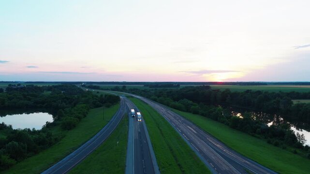 Aerial view of freight trucks hauling cargo along a rural divided highway at dusk, headlights on, with ponds and farm fields fading into a soft sunset. Richview Illinois Above I-64
