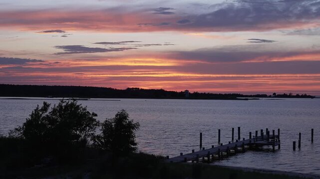 A breathtaking summer sunset over a calm lake featuring a wooden dock silhouetted against vibrant orange, pink, and purple clouds. Peaceful waterfront landscape with reflections on the water, ideal fo