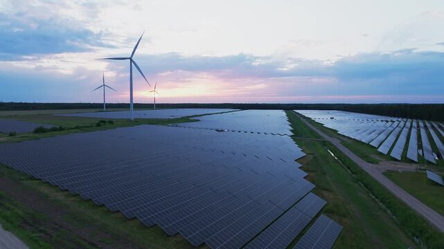Smooth aerial drone footage flying above a large solar power plant with wind turbines during a colorful sunset in Latvia. The video showcases renewable energy infrastructure, photovoltaic panels and c