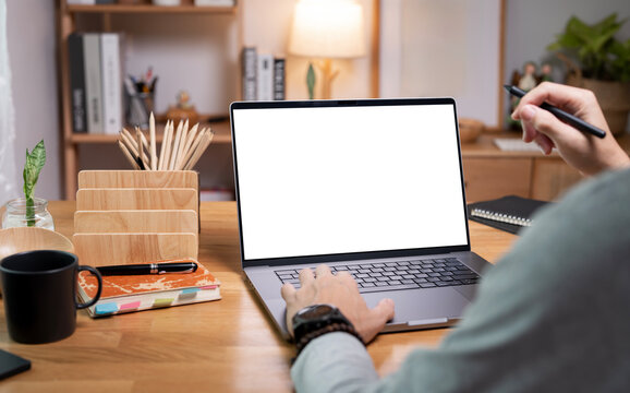 Man hands pointing with stylus at laptop with blank white mockup screen on wooden desk.