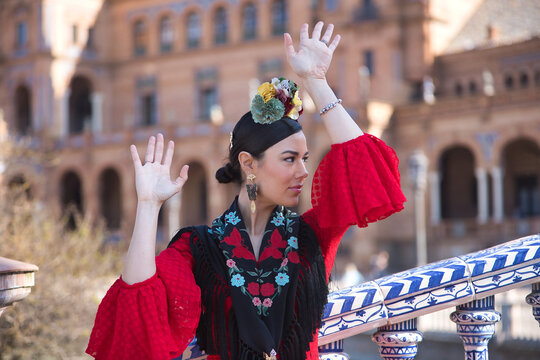 An Andalusian woman wearing a red flamenco dress and a black shawl, with flowers in her hair, dancing flamenco on a beautiful bridge in Seville&rsquo;s Spain Square. Concept of dance, art.