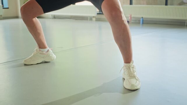 Woman performing jumping jacks and squats in fitness studio, focusing on legs and sneakers. Video is perfect for gym advertisements and workout app promotions